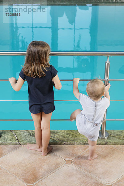 Ein junges Mädchen und ihr kleiner Bruder stehen nebeneinander und schauen in ein Schwimmbad.