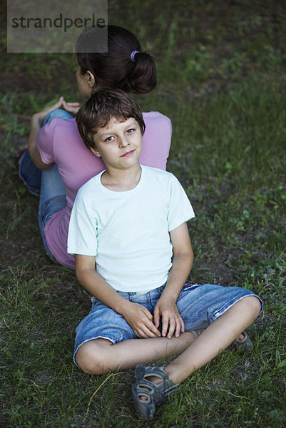 Ein Junge sitzt im Schneidersitz auf Gras und lehnt sich mit dem Rücken gegen den Rücken seiner Mutter.