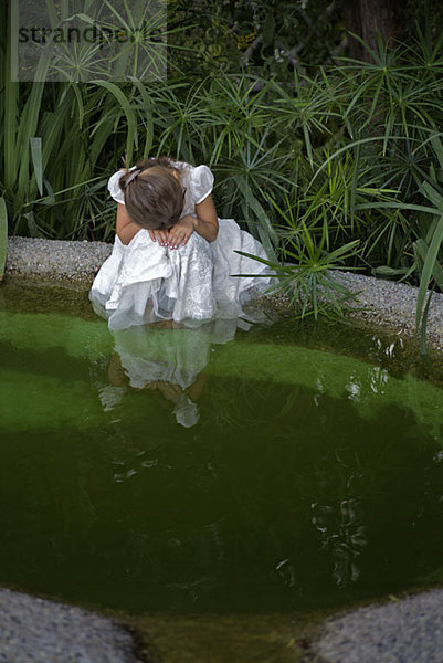 Ein junges trauriges Mädchen  das seinen Kopf auf die Hände legt  während es an einem Teich sitzt.