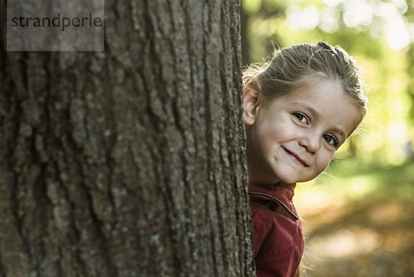 Ein junges lächelndes Mädchen  das hinter einem Baumstamm lugt.