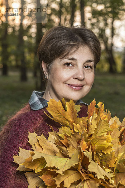 Eine reife Frau  die einen Haufen Herbstblätter in der Natur hält.