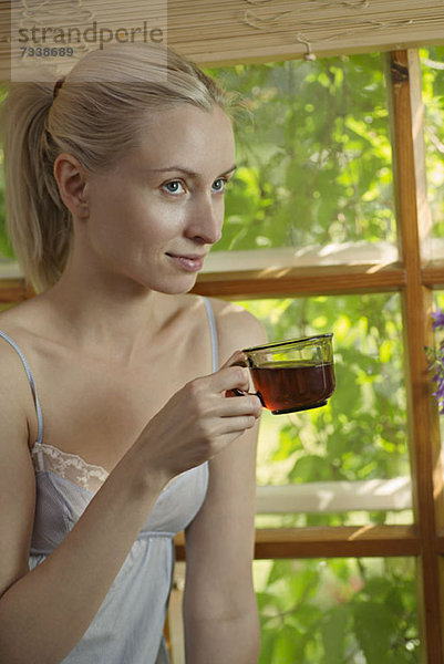 Eine schöne Frau im Nachthemd sitzt am Fenster und trinkt Tee.