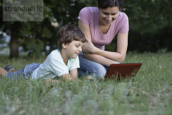 Eine Mutter und ihr Sohn lachen  während sie etwas auf einem Laptop in einem Park sehen.
