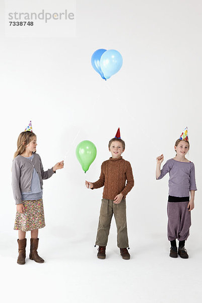 Drei Kinder tragen Partyhüte und halten Luftballons.