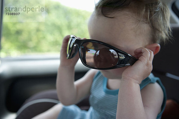 Ein kleiner Junge sitzt in einem stationären Auto und spielt mit einer Sonnenbrille.