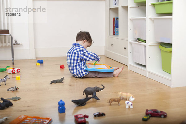 Ein Junge sitzt auf dem Boden und zeichnet auf einer Tafel  umgeben von verschiedenen Spielsachen.