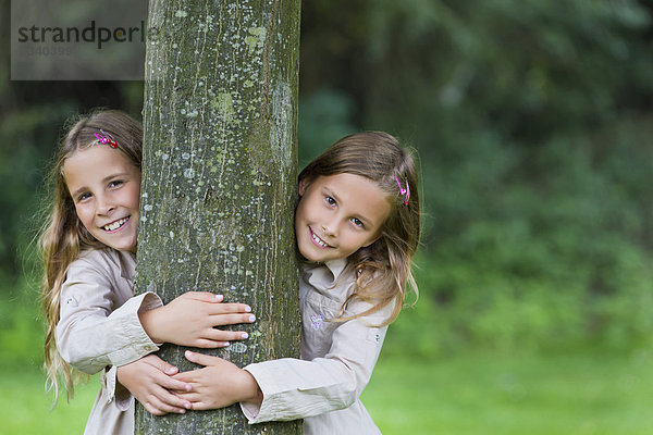 Zwillinge  Mädchen  9 Jahre  schauen links und rechts an einem Baum vorbei