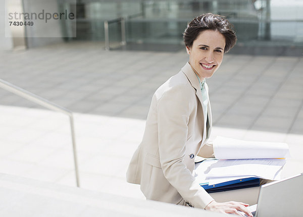 Porträt einer lächelnden Geschäftsfrau mit Laptop und Papierkram außerhalb des Gebäudes