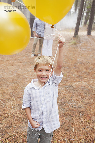 Porträt eines lächelnden Jungen mit Luftballons im Wald und Eltern im Hintergrund