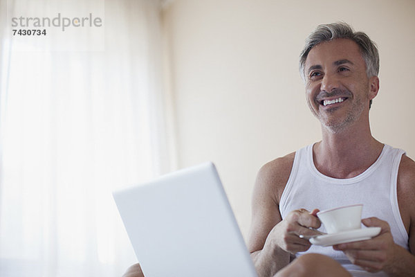 Lächelnder Mann trinkt Kaffee und benutzt Laptop im Schlafzimmer