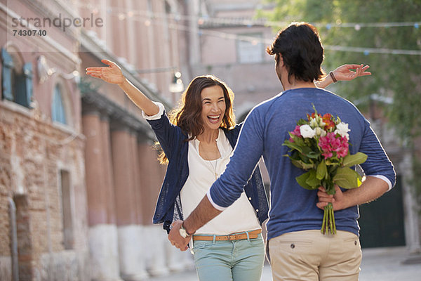 Enthusiastische Frau nähert sich dem Mann mit Blumen hinter dem Rücken.
