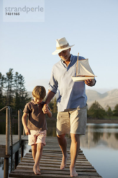 Großvater und Enkel mit Spielzeug-Segelboot beim Spaziergang über den See