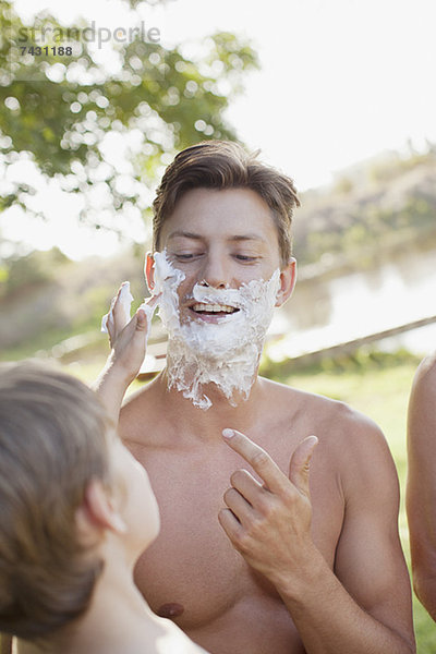 Sohn hilft Vater beim Auftragen der Rasiercreme auf das Gesicht am See.