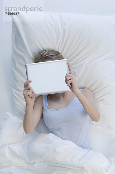 Deutschland  Junge Frau mit digitalem Tablett auf dem Bett liegend
