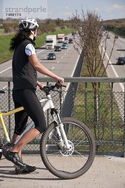 Deutschland  Mittlere erwachsene Frau mit Mountainbike auf Brücke mit Blick auf den Verkehr auf der Autobahn