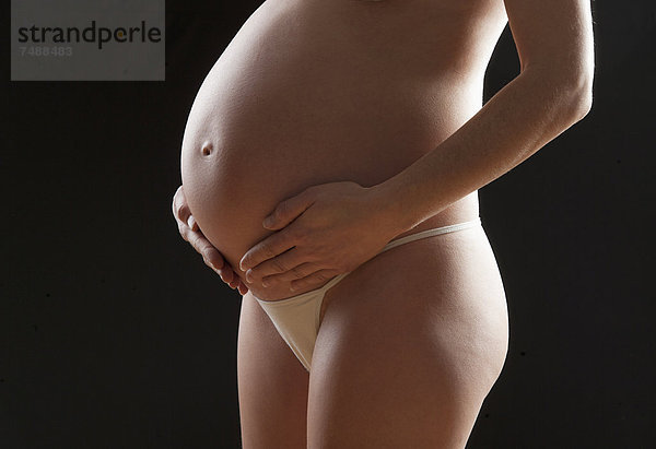 Schwangere Frau berührt ihren Bauch vor schwarzem Hintergrund