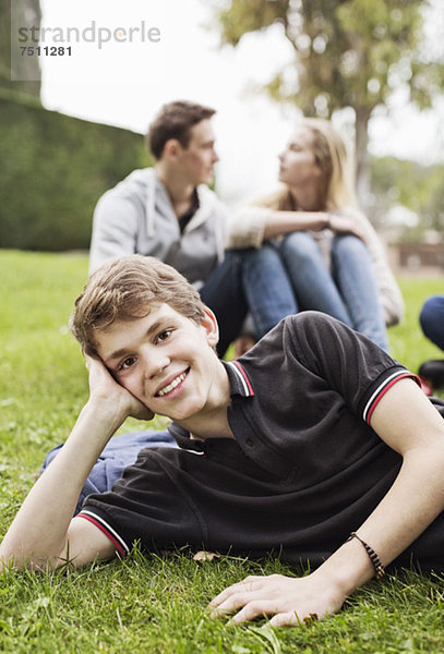 Porträt eines Jungen auf Gras liegend mit einem im Hintergrund sitzenden Paar