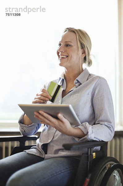 Glückliche behinderte Frau im Rollstuhl mit digitalem Tablett und Kreditkarte im Hintergrund