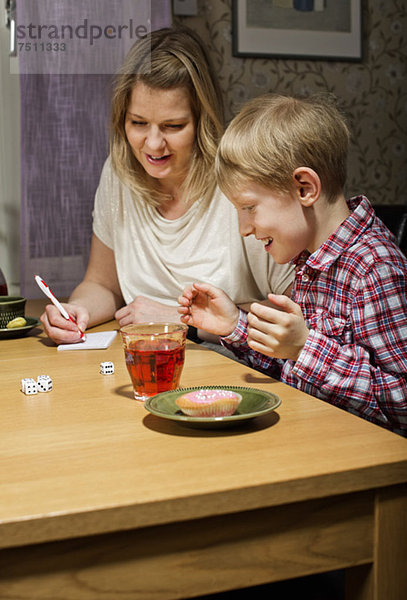 Aufgeregter Junge spielt Würfelspiel  während Mutter am Tisch Partituren schreibt.