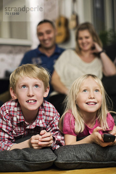 Fröhliche kleine Geschwister beim Fernsehen mit den Eltern im Hintergrund