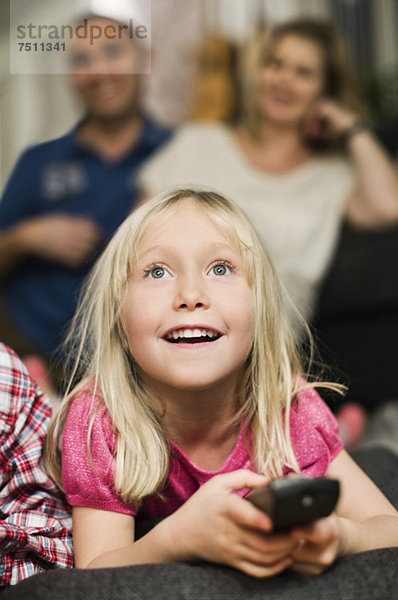 Kleines Mädchen mit Fernbedienung beim Fernsehen und im Hintergrund sitzende Eltern
