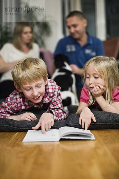 Geschwister lesen Buch im Liegen mit Eltern im Hintergrund