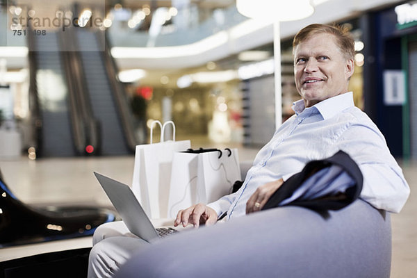 Fröhlicher älterer Mann sitzt auf dem Sofa mit Laptop im Einkaufszentrum