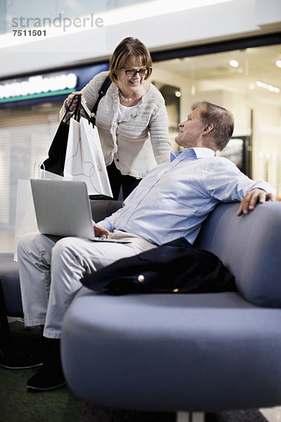 Senior Mann mit Laptop  der Frau mit Einkaufstaschen im Einkaufszentrum ansieht