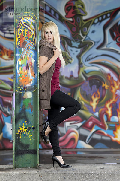 Junge Frau mit langen blonden Haaren posiert an Stahlträger vor Wand mit Graffiti