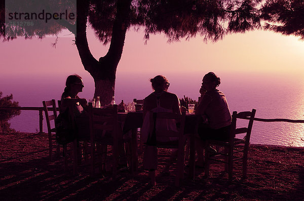 Drei Frauen sitzen bei untergehender Sonne an einem Tisch  Abendstimmung in einem griechischen Garten am Meer  Lefkas  Griechenland  Europa