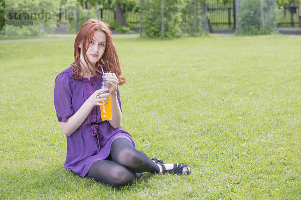Junge Frau sitzt in einem Park und hält eine Flasche Orangenlimonade in der Hand