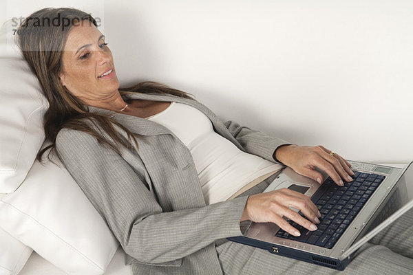 Reife Geschäftsfrau  die auf der Couch liegt und auf dem Laptop tippt.