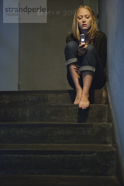 Junge Frau sitzt allein auf der Treppe und schaut auf das Handy.