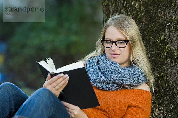 Junge Frau mit Brille sitzt im Park und liest ein Buch