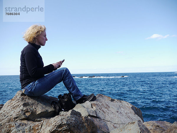 Frau mit Smartphone sitzt auf einem Felsen und schaut aufs Meer  in Roque de las Bodegas  Teneriffa  Kanarische Inseln  Spanien  Europa