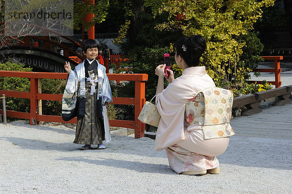 Japanischer Junge in einem dekorativen Kimono  Hakama  Hosenrock und einem Haori  Überjacke  macht Victory-Zeichen  wird von der Mutter in einem Kimono mit großer Obischleife fotografiert  beim Shichi-go-san  Sieben-fünf-drei Fest  Shimogamo Jinja  Kyoto  Japan  Ostasien