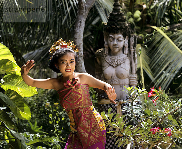 Bali-Tänzerin mit traditionellem Kostüm  vor Steinstatue