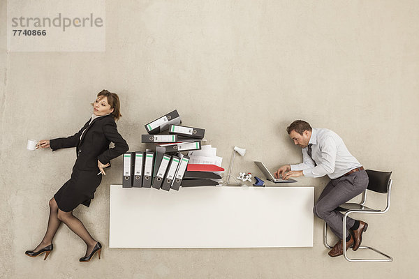 Geschäftsmann  der am Schreibtisch arbeitet  während die Geschäftsfrau sich auf einen Aktenstapel stützt.