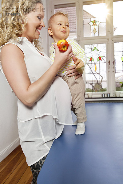 Deutschland  Bonn  Schwangere Mutter isst Apfel mit Sohn im Wohnzimmer  lächelnd