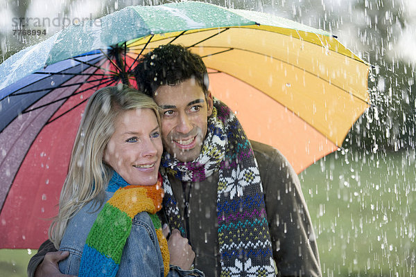 Lächelndes Paar unter regenbogenfarbenem Schirm genießt den Regen