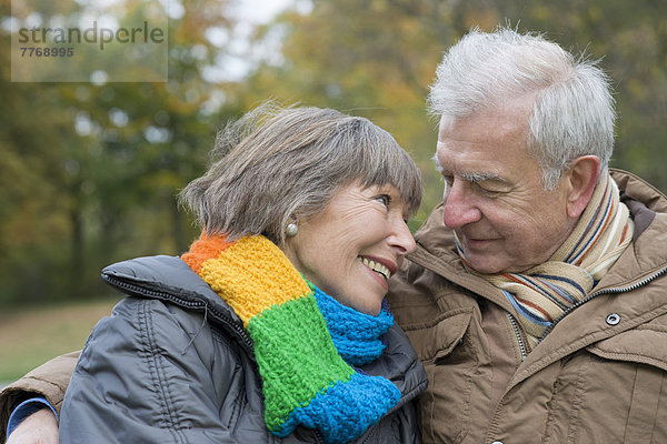 Seniorenpaar sitzt Arm in Arm auf Parkbank und lächelt sich an