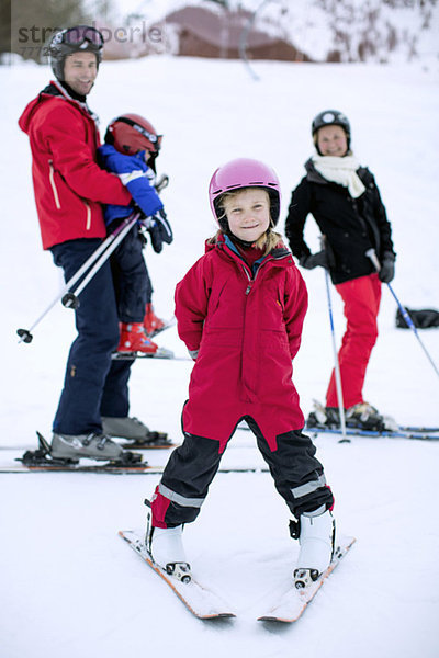 Porträt eines Mädchens in Skikleidung mit lächelnder Familie im Hintergrund