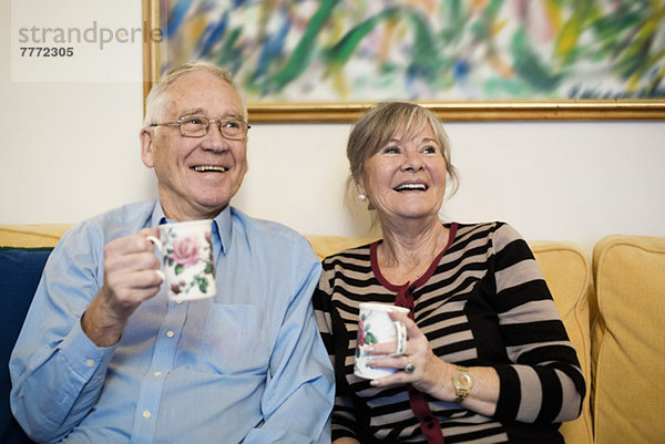 Glückliches Senior-Paar schaut weg und hält Kaffeetassen auf dem Sofa.