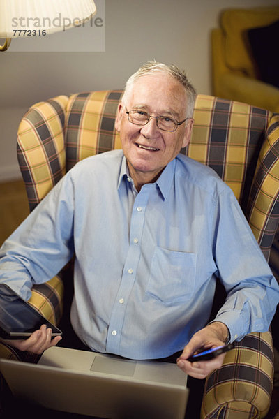 Porträt eines älteren Mannes mit Laptop  Handy und digitalem Tablett auf einem Sessel