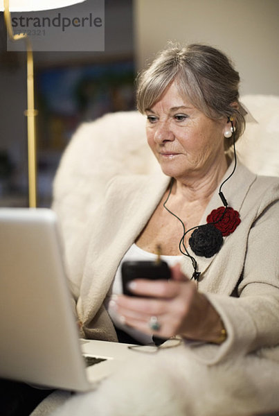 Seniorin mit Laptop beim Musikhören über Handy auf der Couch