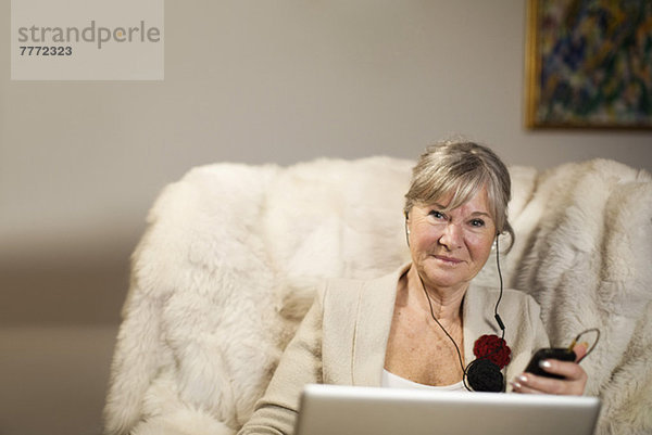 Porträt einer älteren Frau mit Laptop  die Musik hört  während sie auf der Couch sitzt.