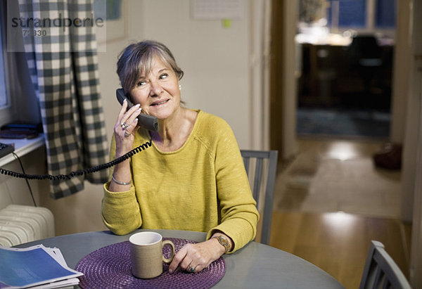 Glückliche Seniorin mit Festnetztelefon am Esstisch