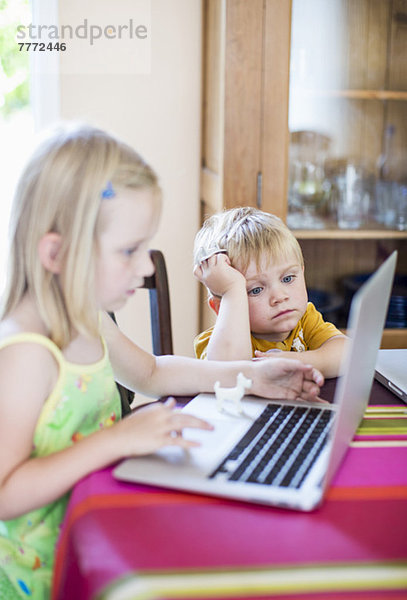 Gelangweilter kleiner Junge  der die Schwester mit dem Laptop ansieht.