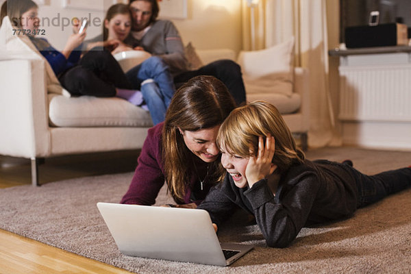 Glückliche Mutter und Sohn mit Laptop auf dem Boden mit Familie auf Sofa im Wohnzimmer sitzen
