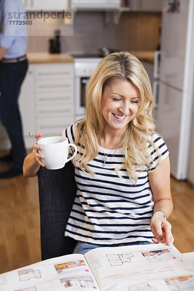 Glückliche Frau mit Kaffeetasse beim Lesen von Immobilienanzeigen in der Zeitung am Frühstückstisch